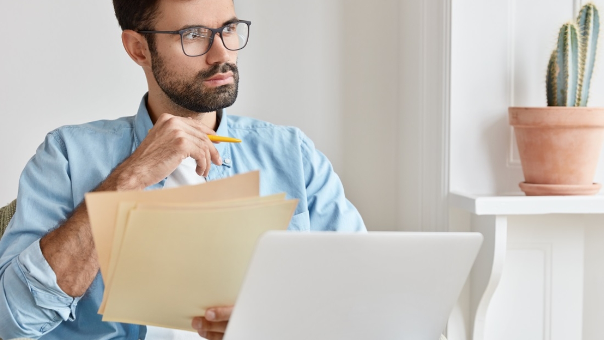 Bearded pensive man works from home, counts financial data, holds paper documents, uses laptop computer for searching information, wears glasses, concentrated aside, develops startup project