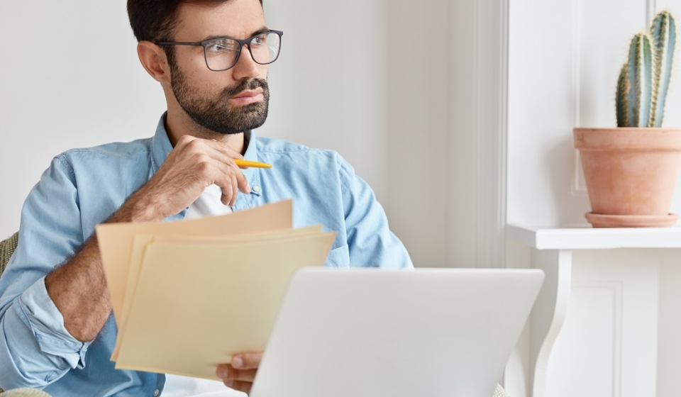 Bearded pensive man works from home, counts financial data, holds paper documents, uses laptop computer for searching information, wears glasses, concentrated aside, develops startup project